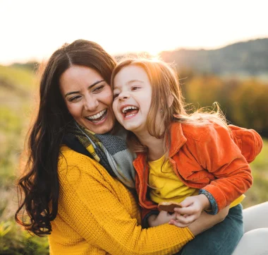 Eine Mutter sitzt mit ihrer kleinen Tochter am Schoß auf einer Wiese, beide lachen und genießen die gemeinsame Zeit.