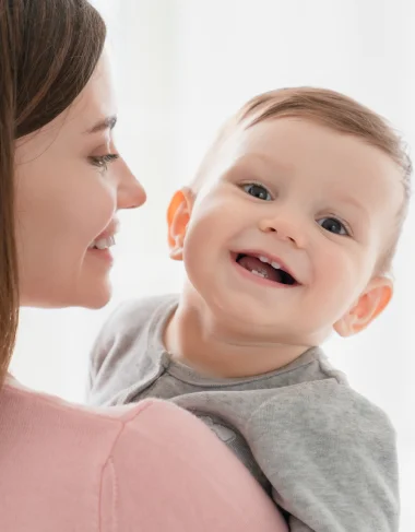 Junge Mutter hat ihr Kleinkind im Arm und lacht es glücklich an.