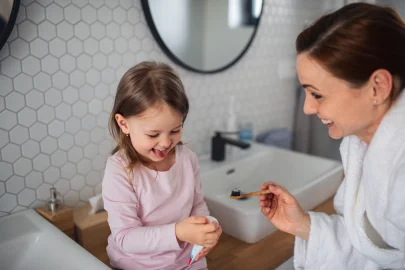 Die Mutter hält die Zahnbürste, während die kleine Tochter versucht, Zahnpasta aus der Tube zu drücken.